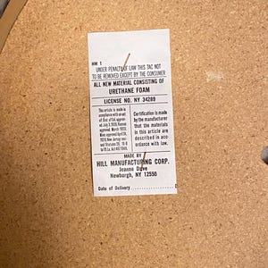 May include: A rectangular white law tag stapled to a light brown particle board surface. The tag contains black printed text stating "UNDER PENALTY OF LAW THIS TAG NOT TO BE REMOVED EXCEPT BY THE CONSUMER" and "ALL NEW MATERIAL CONSISTING OF URETHANE FOAM".