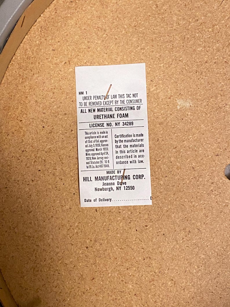 May include: A rectangular white law tag stapled to a light brown particle board surface. The tag contains black printed text stating "UNDER PENALTY OF LAW THIS TAG NOT TO BE REMOVED EXCEPT BY THE CONSUMER" and "ALL NEW MATERIAL CONSISTING OF URETHANE FOAM".