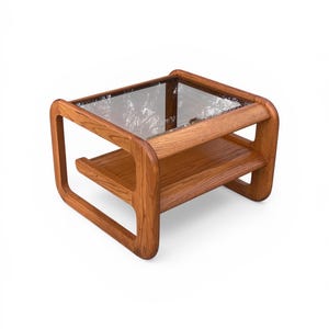 May include: A mid-century modern coffee table with a clear glass top and a lower shelf. The table is constructed from light brown wood and features rounded edges and a rectangular shape. The dimensions are approximately 24 inches long, 24 inches wide, and 18 inches high.