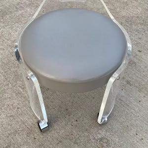 May include: A round stool featuring a gray upholstered seat cushion supported by a clear acrylic frame. The frame includes an integrated handle at the top and is mounted on four small wheels with metallic accents at the base of each leg.