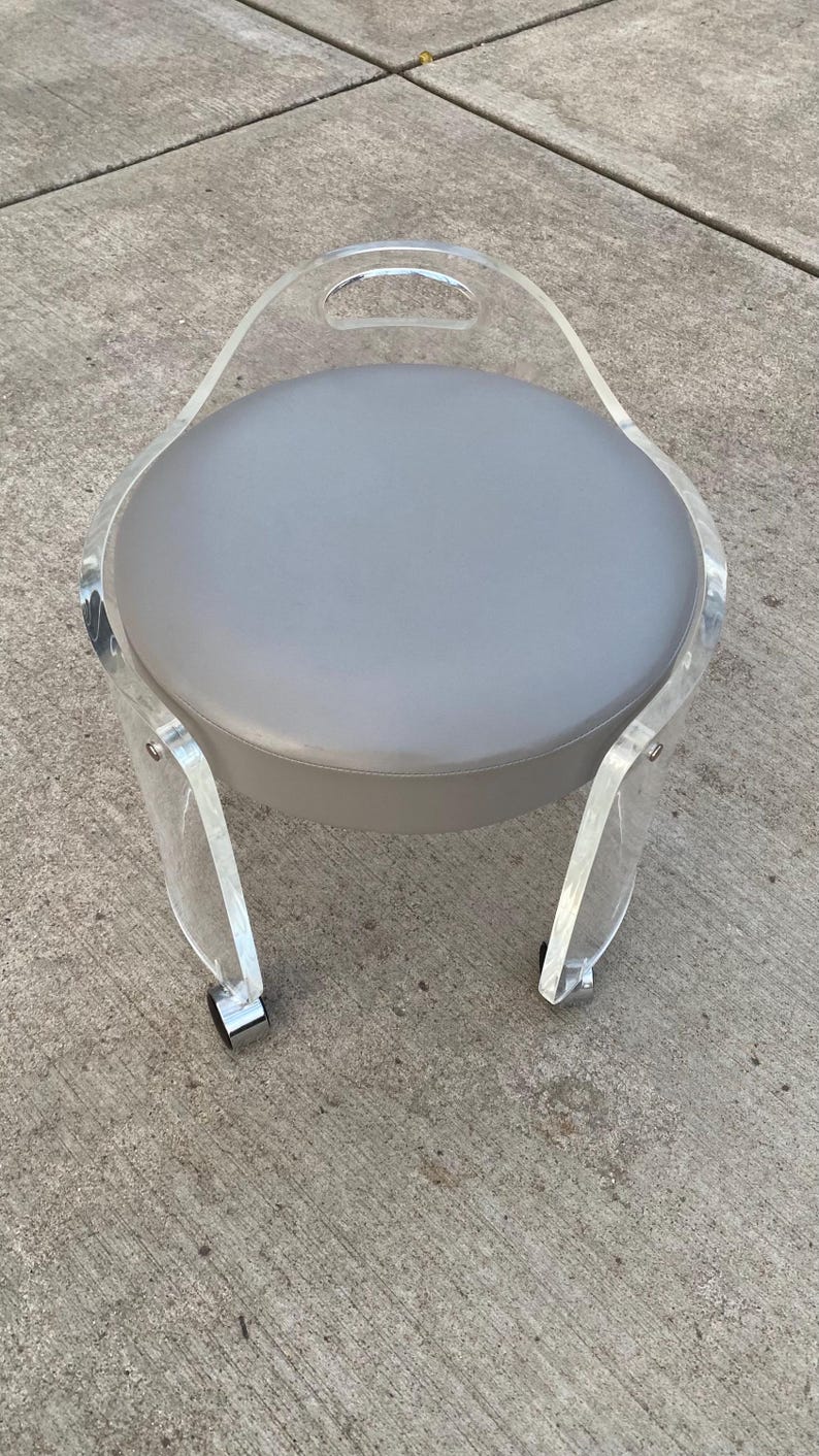 May include: A round stool featuring a gray upholstered seat cushion supported by a clear acrylic frame. The frame includes an integrated handle at the top and is mounted on four small wheels with metallic accents at the base of each leg.
