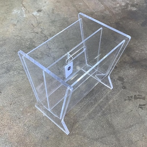 May include: A clear acrylic magazine rack with a simple design. The rack is made of two pieces of acrylic that are joined together to form a rectangular shape. The rack is designed to hold magazines or other items in an upright position.