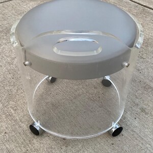 May include: A round stool featuring a clear acrylic cylindrical base with a built-in handle and a smooth gray padded seat. The base is supported by four small wheels with metallic accents, providing mobility to this circular seating piece.