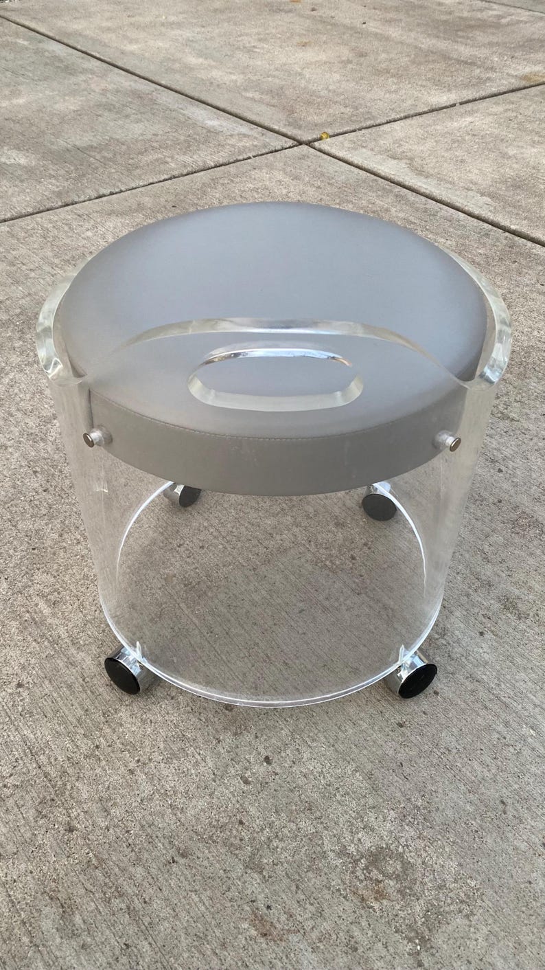 May include: A round stool featuring a clear acrylic cylindrical base with a built-in handle and a smooth gray padded seat. The base is supported by four small wheels with metallic accents, providing mobility to this circular seating piece.