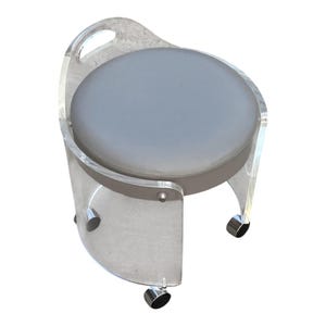 May include: A round stool featuring a clear acrylic frame with a curved backrest and a handle cutout. The seat is upholstered in a smooth, light gray material. The base is supported by four small, metallic swivel casters for mobility.