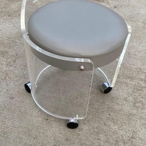 May include: A rolling stool featuring a clear acrylic frame with a curved backrest and a circular seat upholstered in smooth gray material. The base is supported by three black casters, and the structure includes a metal fastener on the side.