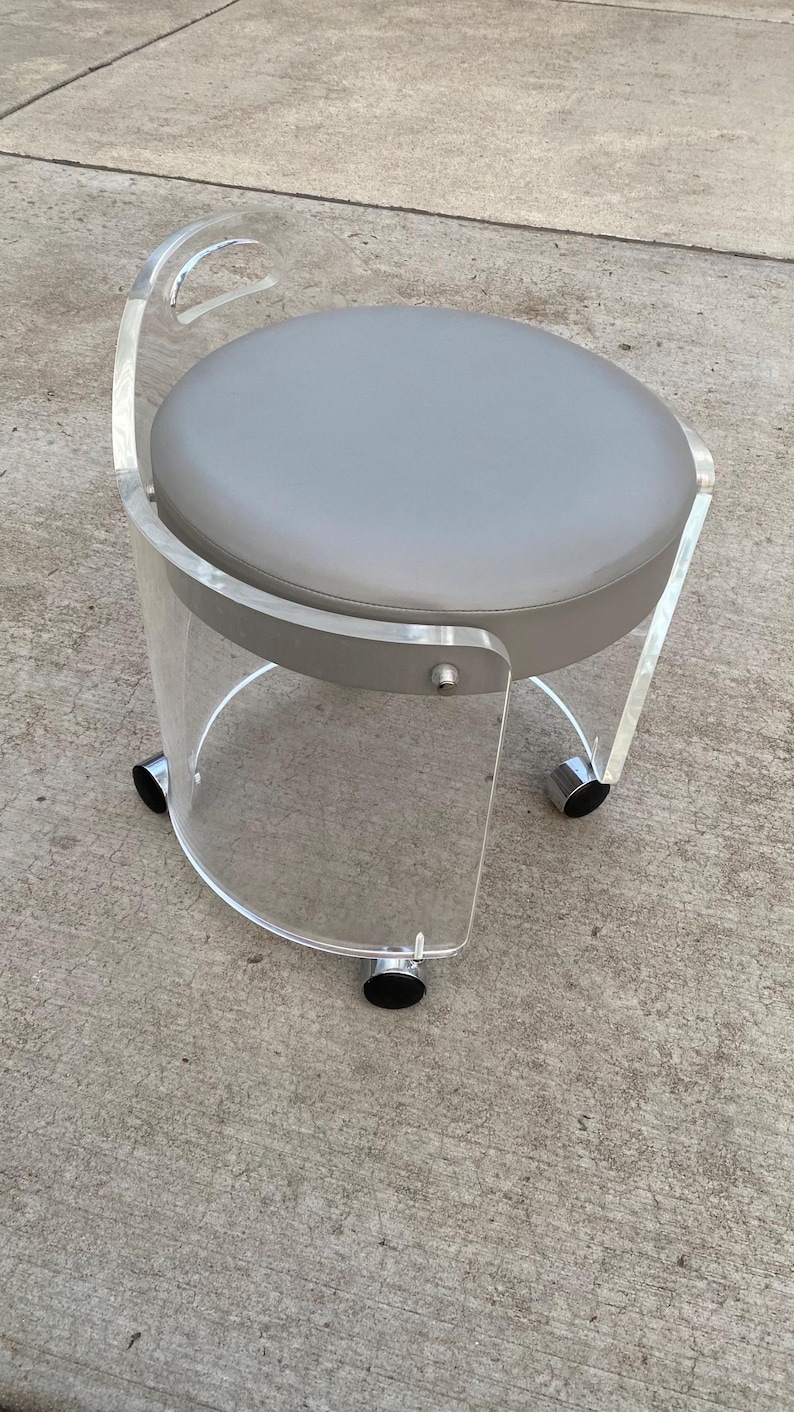 May include: A rolling stool featuring a clear acrylic frame with a curved backrest and a circular seat upholstered in smooth gray material. The base is supported by three black casters, and the structure includes a metal fastener on the side.