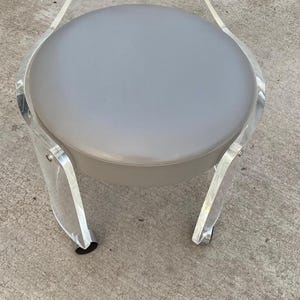 May include: A round stool featuring a clear acrylic frame with a curved backrest and a handle cutout. The seat is upholstered in a smooth, solid light gray material. The structure is supported by four curved acrylic legs with small casters.