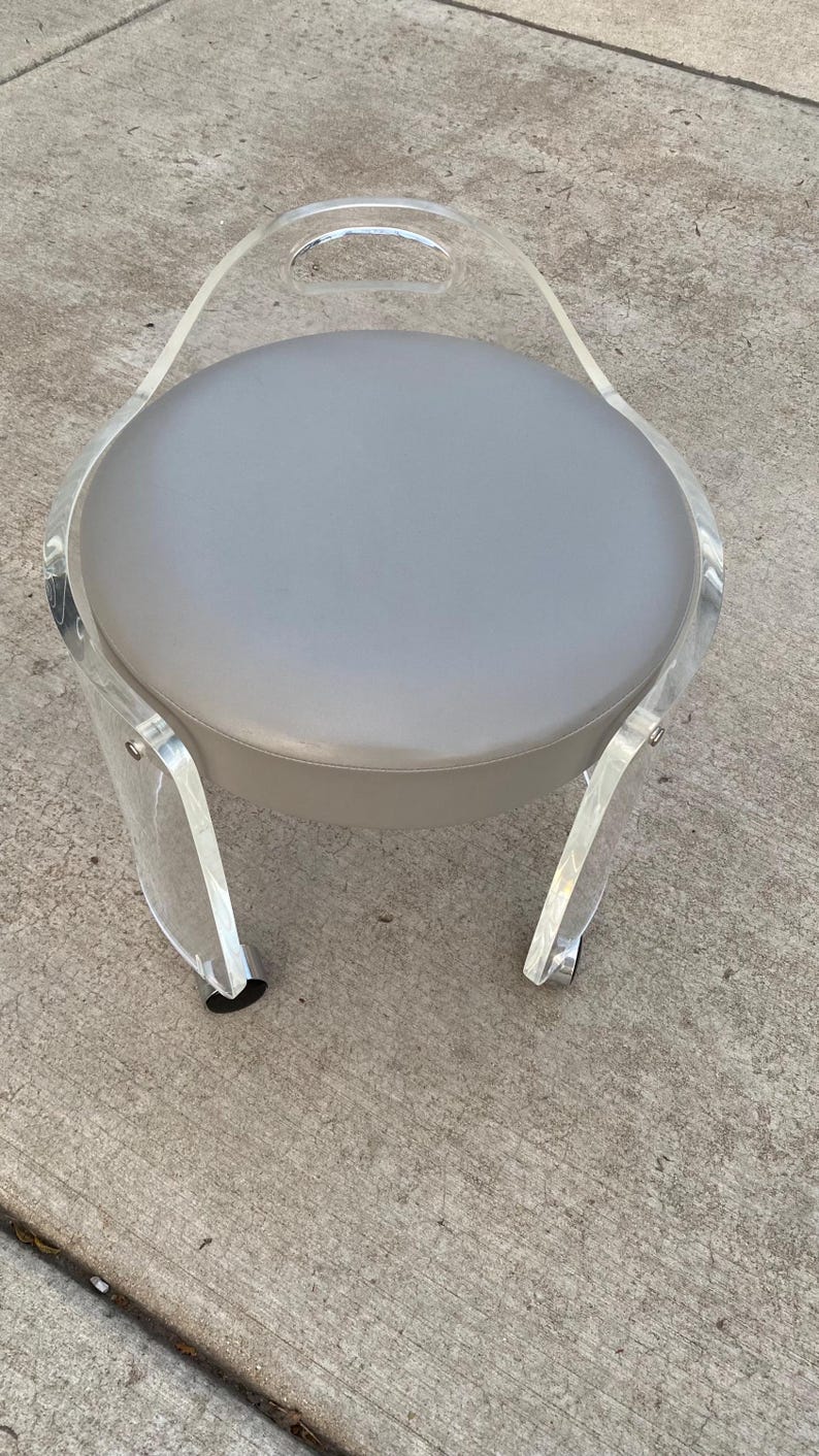 May include: A round stool featuring a clear acrylic frame with a curved backrest and a handle cutout. The seat is upholstered in a smooth, solid light gray material. The structure is supported by four curved acrylic legs with small casters.