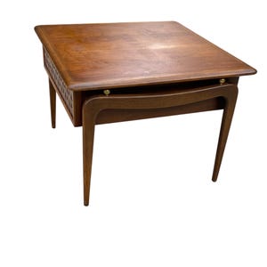 May include: A mid-century modern side table with a square top and a lower shelf. The table is made of wood with a warm brown finish. The legs are tapered and angled, and the table has a drawer with a decorative carved design.