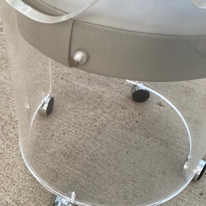 May include: A round stool featuring a clear acrylic cylindrical base and a padded light gray seat. The base is supported by four small silver-toned casters with black wheels, and a single silver-toned metal button is visible on the side of the seat.