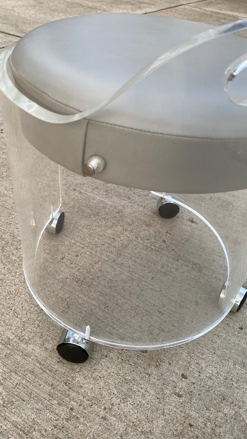 May include: A round stool featuring a clear acrylic cylindrical base and a padded light gray seat. The base is supported by four small silver-toned casters with black wheels, and a single silver-toned metal button is visible on the side of the seat.