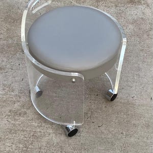 May include: A round stool featuring a clear acrylic frame with a curved backrest and a circular gray padded seat. The base is supported by four silver-toned metal casters, allowing for mobility. The design incorporates a handle cutout in the back.