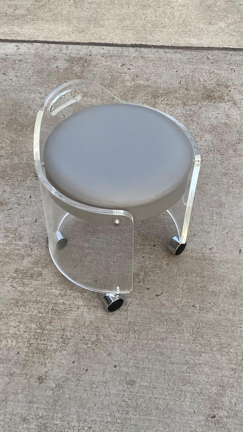May include: A round stool featuring a clear acrylic frame with a curved backrest and a circular gray padded seat. The base is supported by four silver-toned metal casters, allowing for mobility. The design incorporates a handle cutout in the back.