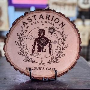 May include: A round wooden plaque with a dark brown bark edge. The plaque features the text "Astarion Girl Dinner" and "Baldur's Gate" with a detailed illustration of a figure, sun, stars, and leaves. The plaque is displayed on a black metal stand.
