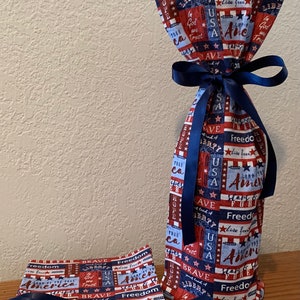 May include: Red, white, and blue fabric wine bottle bag with a navy blue ribbon. The bag is decorated with stars, stripes, and patriotic phrases like "Freedom", "Brave", "Liberty", and "America".