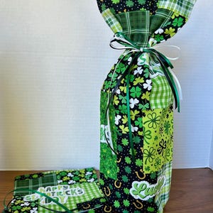 May include: A patchwork wine bottle bag in shades of green, black, and white, featuring clover and horseshoe patterns. The bag is tied with green and white ribbons. A matching coaster and ribbon are also visible, with the text "Happy St. Patrick's Day".