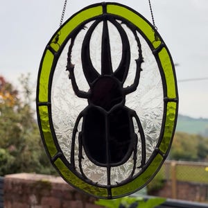 May include: An oval stained glass window hanging with a black beetle design. The beetle is set within a clear, textured glass pane, framed by a bright green border. A decorative chain is attached for hanging.