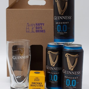 May include: A brown cardboard box with a handle, containing four cans of Guinness 0.0% alcohol-free stout, a glass pint glass, and a small yellow box of Lancashire cheese and spring onion biscuits. The box has a purple logo that says "Happy Days Drinks".