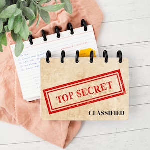 May include: A small disc-bound notebook with a tan cover featuring a red rectangular stamp that reads TOP SECRET and the word CLASSIFIED in black text. The notebook is open to a lined page with handwritten notes and a yellow sticky note attached.