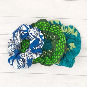 May include: Three fabric hair scrunchies featuring distinct patterns. One is blue and white with a geometric design, one is dark green with a bright green diamond grid, and one is a teal and light green abstract print with a soft, gathered texture.