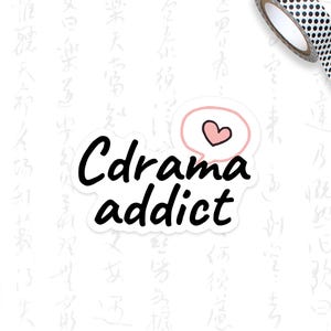 May include: A white sticker with the text "Cdrama addict" in a black, bold font. A pink speech bubble with a heart is above the word "addict". A roll of black and white polka dot washi tape and a light green pencil are in the background.