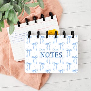 May include: A small spiral-bound notebook with a white cover featuring a repeating pattern of light blue bows and small dots. The word NOTES is printed in dark blue capital letters in the center. The notebook has black plastic binding rings.