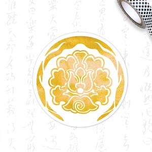May include: A round, white sticker with a gold floral design. The design features a stylized flower within a gold border. A roll of black and white polka dot washi tape and a light green pencil are in the background.
