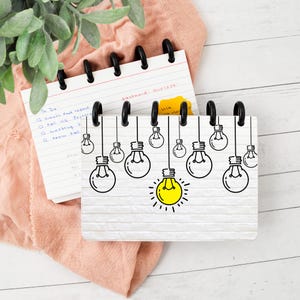 May include: A small spiral-bound notebook with a white brick wall pattern on the cover, featuring a black line art illustration of multiple hanging light bulbs, with one central bulb highlighted in bright yellow, and black plastic binding rings.