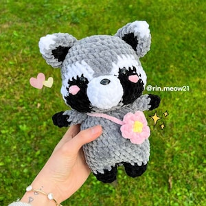 May include: A gray and black crocheted raccoon plush toy with a pink flower on its shoulder. The toy has a white face with black eyes and pink cheeks. The toy is being held in a hand with a gold bracelet. The background is green grass.