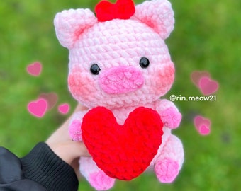 Könnte beinhalten: Ein rosa gehäkeltes Schwein, das ein rotes gehäkeltes Herz hält. Das Schwein hat eine rote Schleife auf dem Kopf und rosafarbene Wangen. Das Schwein ist von verschwommenen rosa Herzen umgeben.