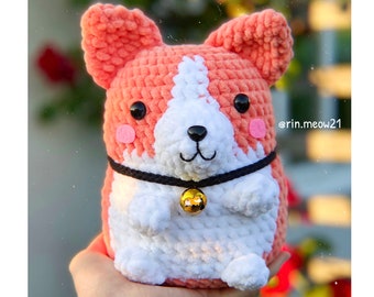 May include: A crocheted plush toy shaped like a corgi dog. The dog is wearing a black collar with a gold bell. The dog is pink and white with black eyes and a black nose.