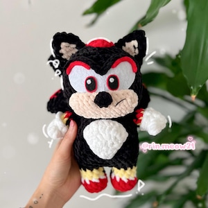 Może przedstawiać: Ręcznie robiona czarno-czerwono-biała pluszowa zabawka z szydełka. Plusz przedstawia postać z czarnym korpusem, czerwonymi akcentami, białą twarzą i dłońmi oraz czerwono-żółtymi stopami. Zabawka jest trzymana na tle zielonego, liściastego tła.