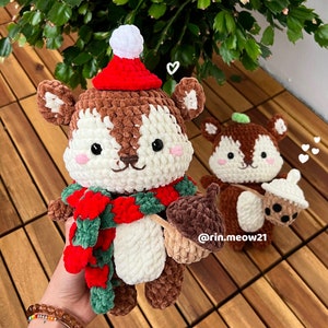 Puede incluir: Dos peluches de ardilla de crochet, uno con un gorro de Papá Noel rojo y una bufanda verde, sosteniendo una bellota marrón, y el otro sosteniendo una taza de té boba marrón. Las ardillas tienen pelaje marrón y blanco y ojos negros.