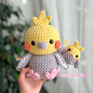 May include: A handmade crochet bird plushie in yellow, grey, and pink. The bird has a yellow crest, orange cheek patches, and black eyes. A smaller bird is attached to the larger one. The plushie is held in a hand.