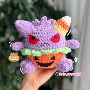 May include: A handmade plush toy in the shape of a purple creature with red eyes and a candy corn horn. The creature is wearing an orange pumpkin costume with a carved face and green trim. The toy is held in a hand.