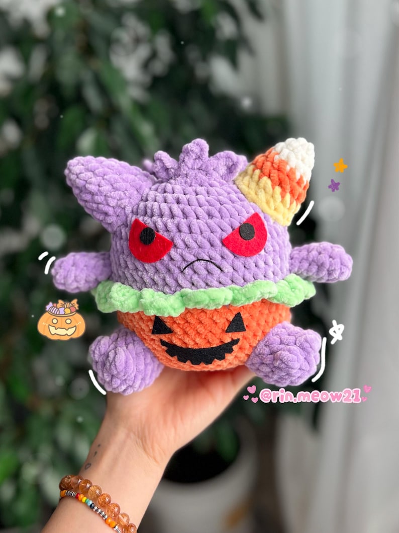 May include: A handmade plush toy in the shape of a purple creature with red eyes and a candy corn horn. The creature is wearing an orange pumpkin costume with a carved face and green trim. The toy is held in a hand.