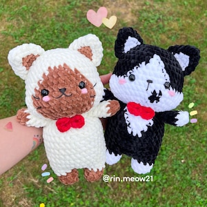 May include: Two crocheted plush toys shaped like cats. One cat is white with brown fur on its face and a red bow tie. The other cat is black with white fur on its face and a red bow tie. The cats are sitting on a green grassy surface. The text "@rin.meow21" is visible in the bottom right corner of the image.