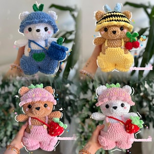 May include: Four handmade crochet plush bears wearing hats and overalls. One white bear in blue, one brown bear in yellow with a bee theme, one brown bear in pink with a strawberry theme, and one white bear in pink with a strawberry theme.
