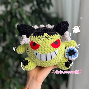 May include: A handmade, lime green crochet plush toy with black hair, gray accents, and red eyes. The toy has a stitched mouth with white teeth and a small blue and white ball. The toy is held in a hand.