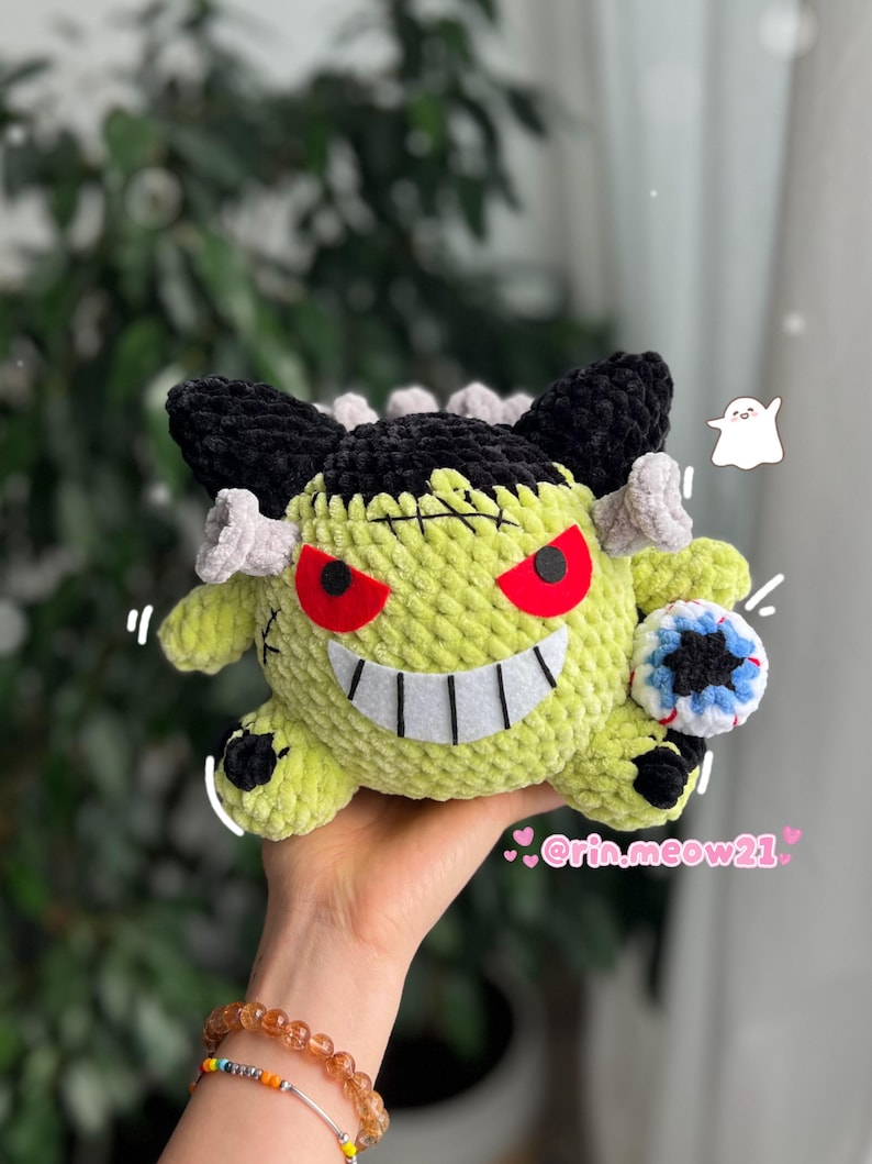 May include: A handmade, lime green crochet plush toy with black hair, gray accents, and red eyes. The toy has a stitched mouth with white teeth and a small blue and white ball. The toy is held in a hand.