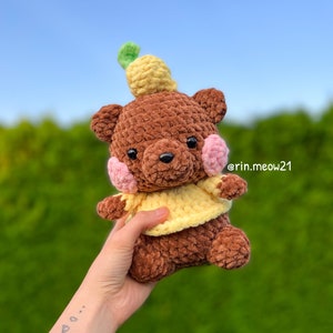 May include: A brown crocheted teddy bear wearing a yellow and pink crocheted outfit with a yellow crocheted pineapple on its head. The bear is being held by a hand with a small tattoo on the wrist.