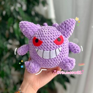 May include: A handmade, plush, lavender-colored Gengar, a character from the Pok&eacute;mon series, with red eyes and a wide, toothy grin. The plush has small arms and ears. The image includes the Instagram handle @rin.meow21.