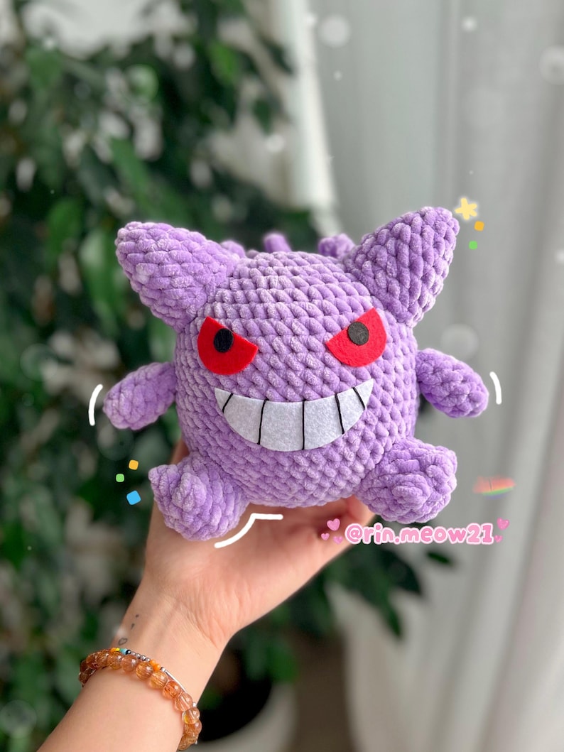 May include: A handmade, plush, lavender-colored Gengar, a character from the Pok&eacute;mon series, with red eyes and a wide, toothy grin. The plush has small arms and ears. The image includes the Instagram handle @rin.meow21.