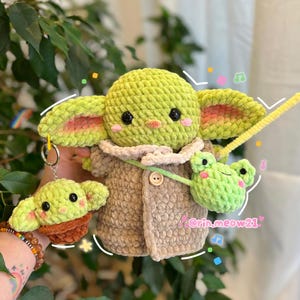 May include: A green crocheted plush toy with a brown crocheted coat and a green crocheted frog keychain. The toy has large eyes and a small mouth. The frog keychain has a small mouth and large eyes. The toy is sitting on a person's arm.