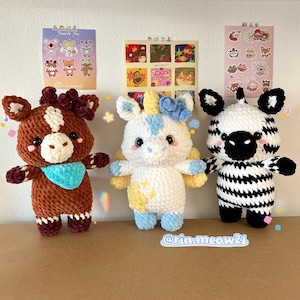 Può includere: Tre animali di peluche all'uncinetto: un cavallo marrone con un bavaglino blu, un unicorno bianco con accenti gialli e blu e una zebra in bianco e nero. Gli animali sono tutti in piedi su una superficie marrone.