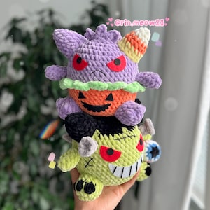 May include: Three handmade crochet plush toys stacked on top of each other. The top plush is purple with red eyes and a candy corn-colored horn. The middle plush is an orange pumpkin. The bottom plush is green with black hair and a stitched mouth.