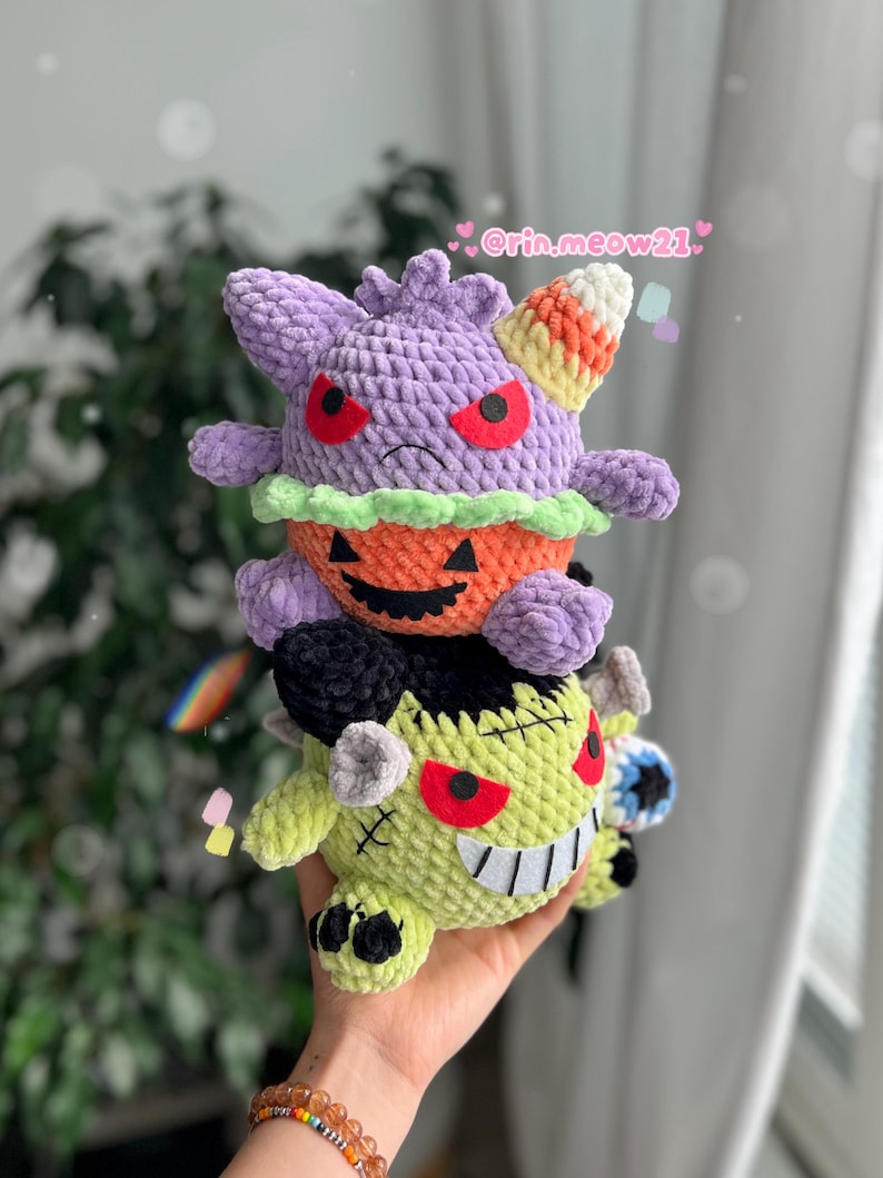 May include: Three handmade crochet plush toys stacked on top of each other. The top plush is purple with red eyes and a candy corn-colored horn. The middle plush is an orange pumpkin. The bottom plush is green with black hair and a stitched mouth.