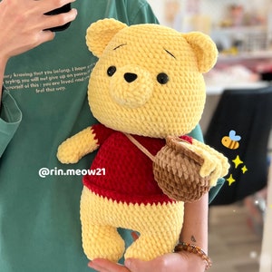 May include: A crocheted Winnie the Pooh plush toy wearing a red shirt and carrying a brown honey pot. The toy is yellow with a black nose and brown eyes. The background is a person wearing a green shirt.