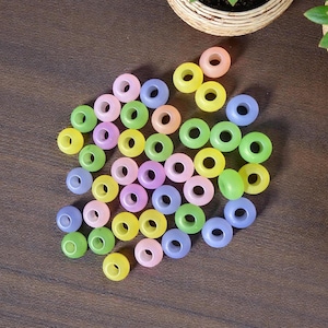 May include: A collection of colorful, translucent donut-shaped beads in shades of pink, yellow, green, and blue. The beads are scattered on a dark brown wooden surface, suggesting a craft or jewelry-making theme.